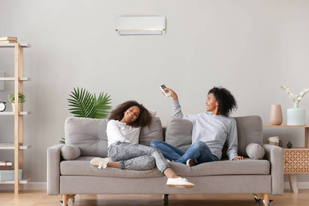 Happy family relax on sofa under air conditioner, black mom holding remote control switch on conditioning in living room adjust comfort temperature for daughter, climate system at modern home