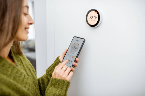Woman dressed in green sweater regulating heating temperature with a modern smart thermostat and smart phone at home. Synchronization of thermostat with mobile devices concept